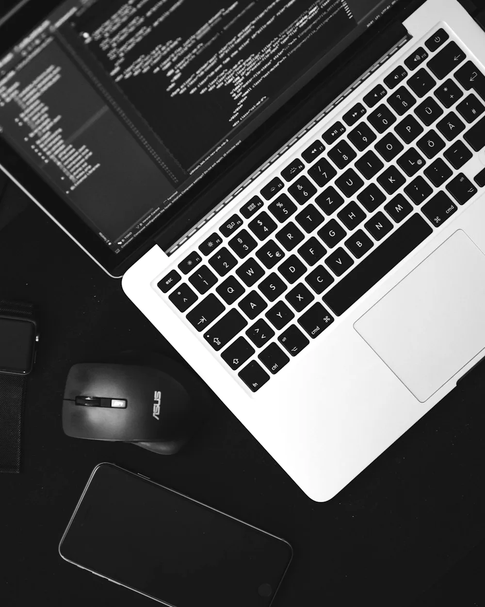 A MacBook open on a code editor, on a dark desk beside a mouse and phone